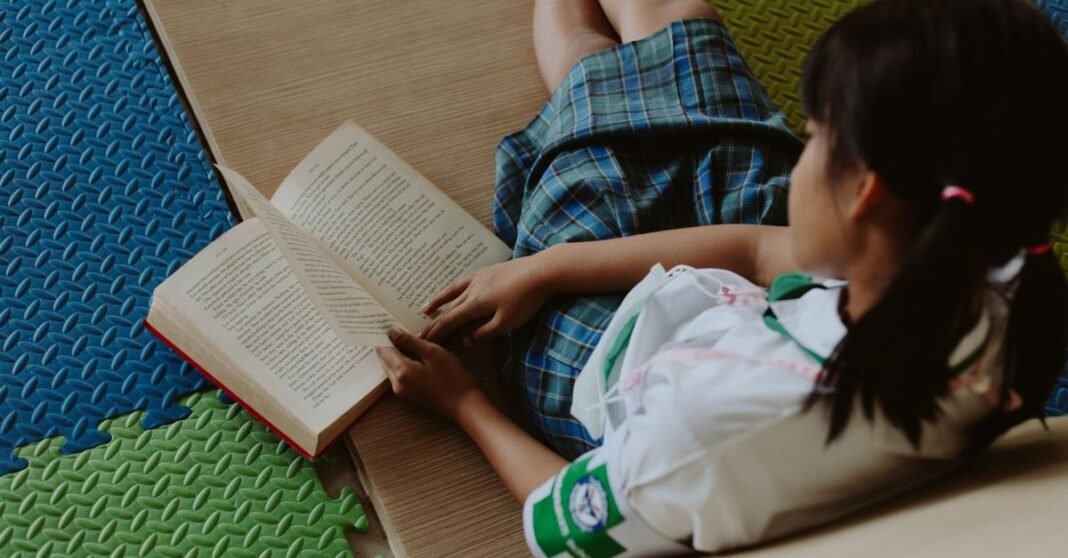 A young girl in a school uniform reading a book while sitting on a colorful mat indoors.