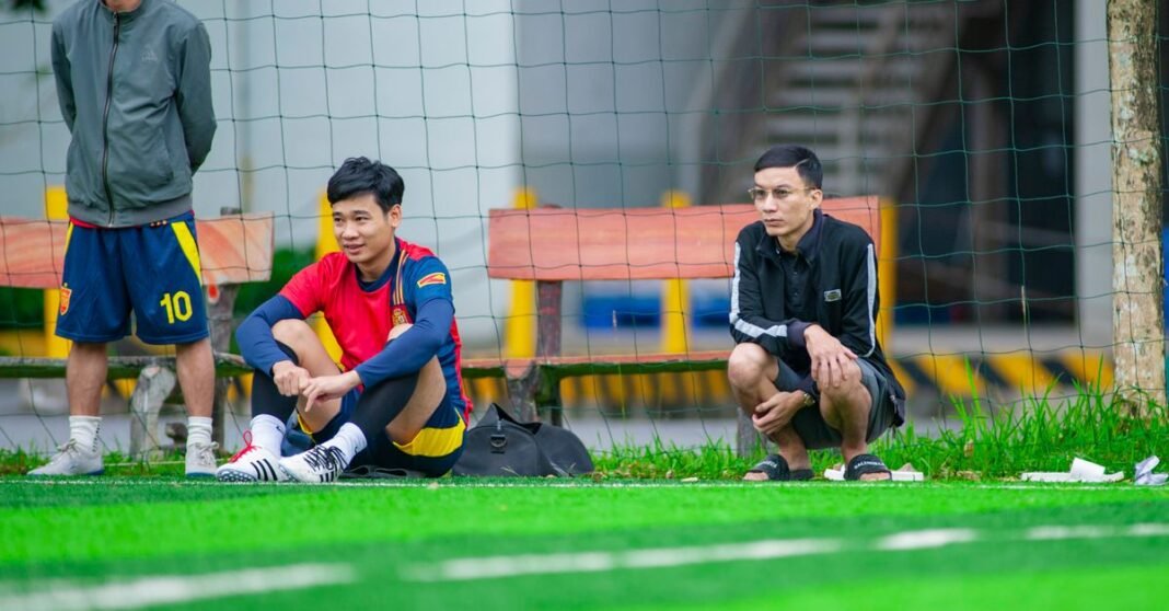 Football players sitting on the sideline of a green field in Hanoi, Vietnam.