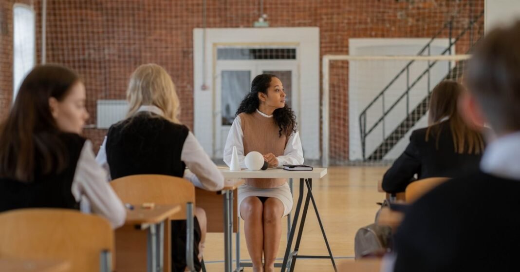 Teacher sitting at desk during class in a school gymnasium.