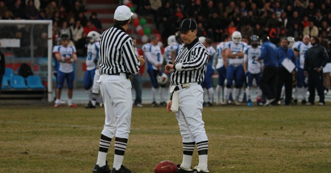 Two referees on a football field preparing for a game with players in the background at a stadium.