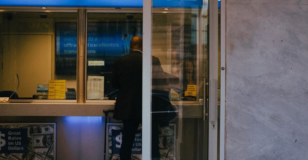 Man at a currency exchange office window, showing currency rates inside a bustling city.