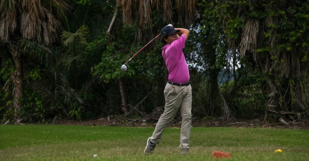 Adult male golfer swinging a club on a lush, green golf course surrounded by trees.