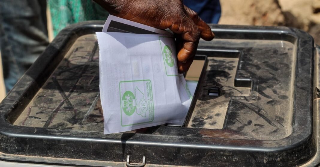 A close-up shot of a hand placing a paper ballot into a transparent voting box outdoors.