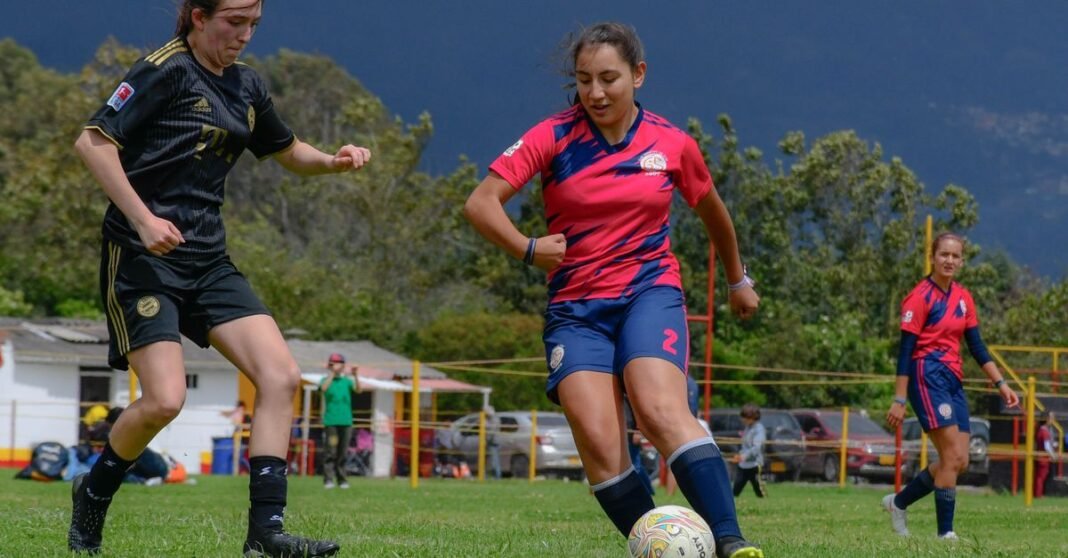 Women soccer players in action during a match on a lush green field with a mountain backdrop.