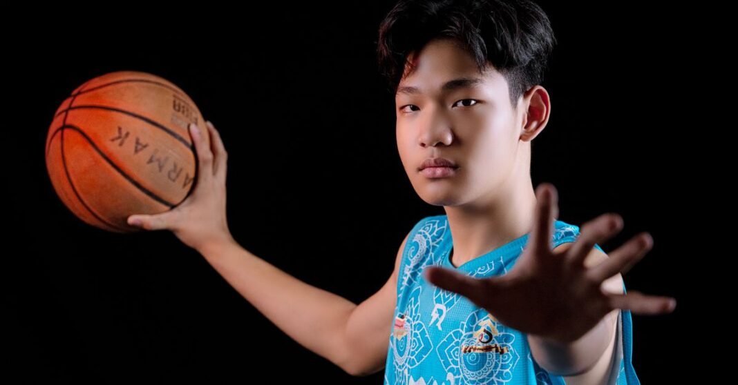 Teenager in blue jersey posing with a basketball against black backdrop.