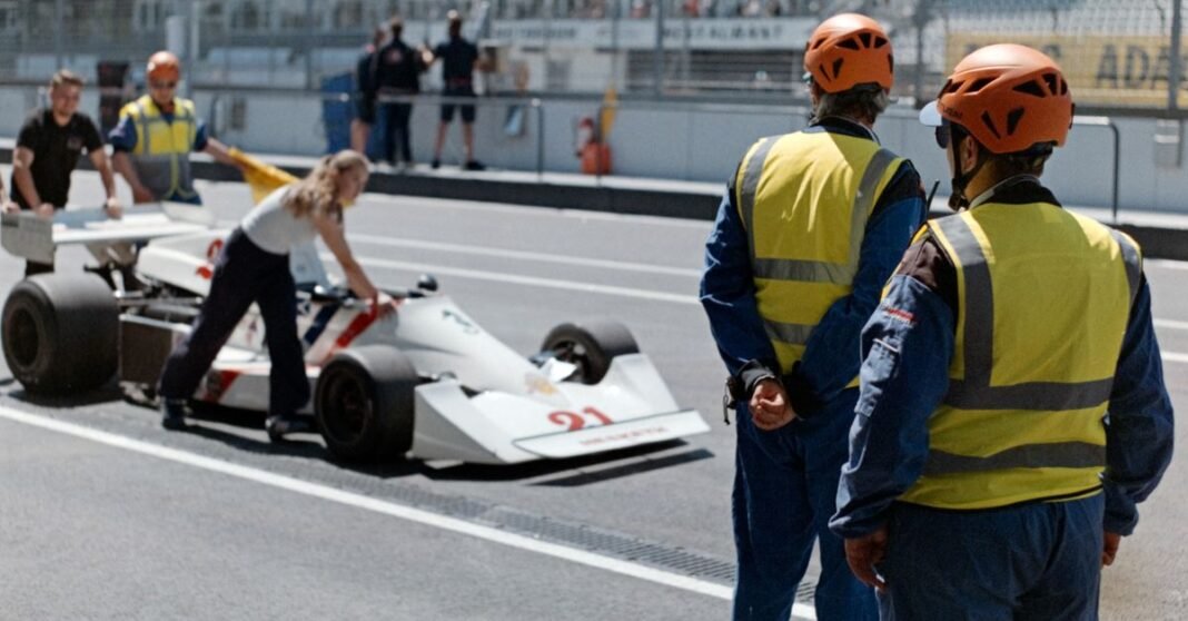 Race car being wheeled to pit stop with crew in helmets and safety vests.