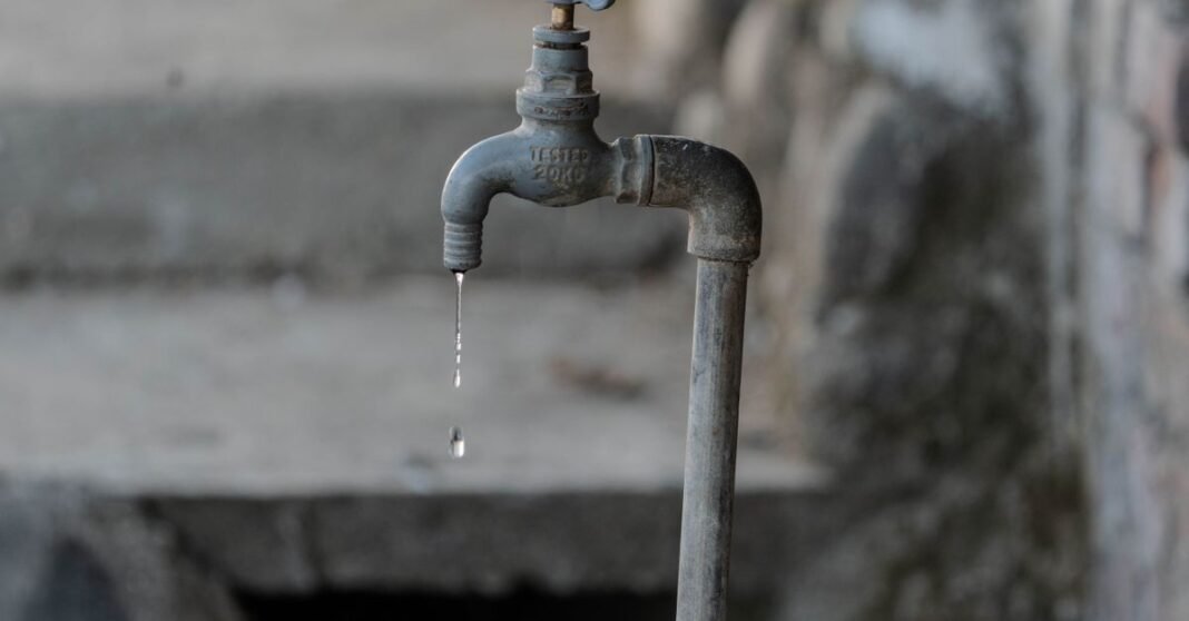 A close-up of a rustic outdoor faucet dripping water against a natural, textured background, symbolizing scarcity.