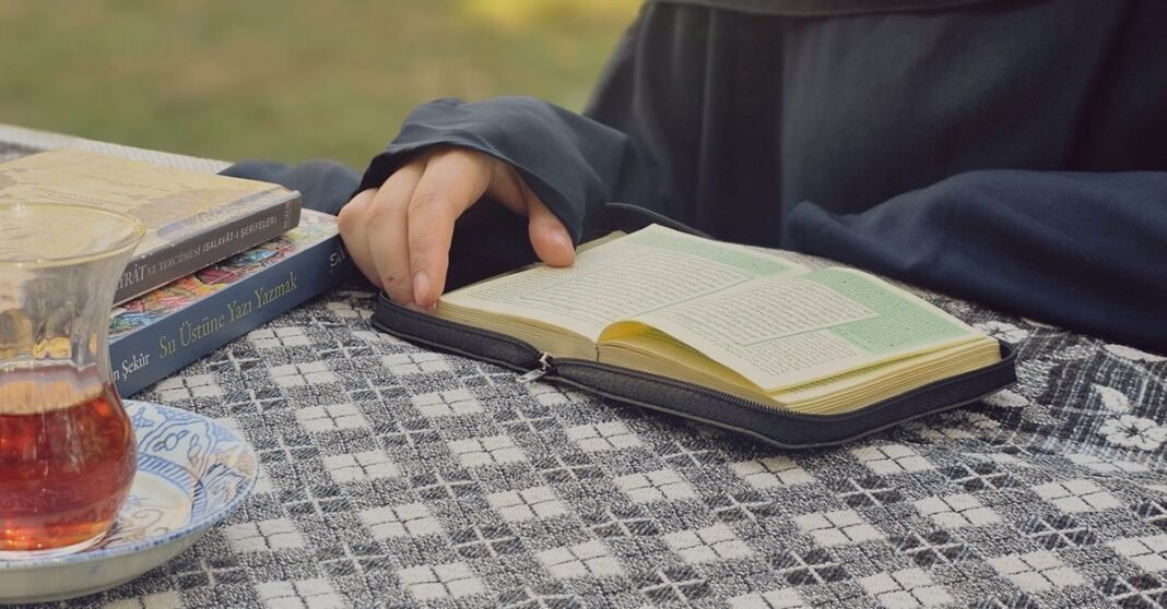 A person reading a book outdoors with a glass of tea on a patterned tablecloth.