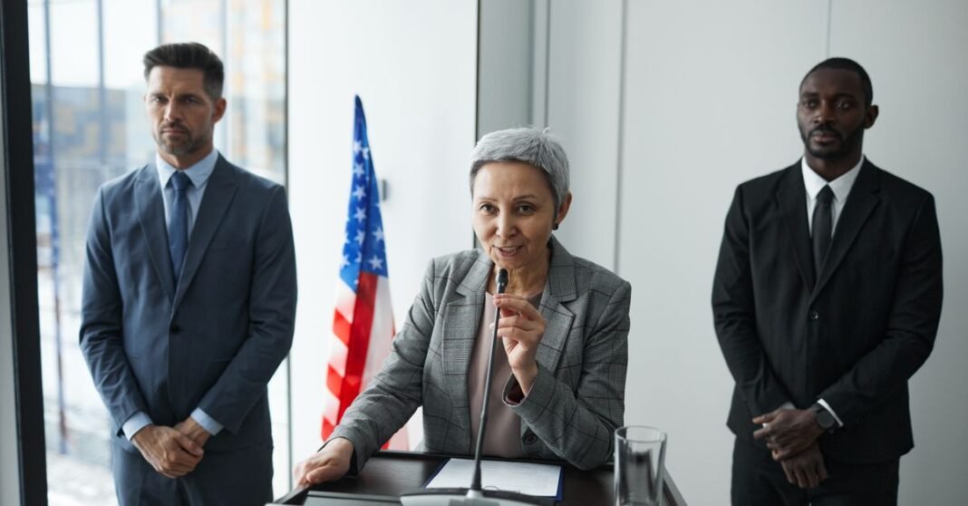 A female politician delivers a speech with bodyguards and an American flag in the background.