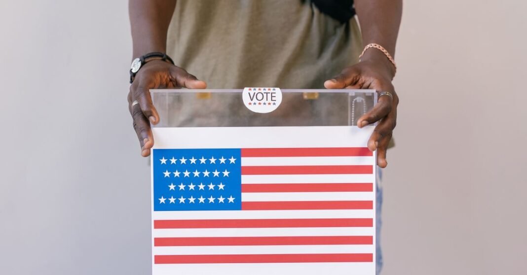 Close-up of a person placing a vote in a transparent ballot box with an American flag print.
