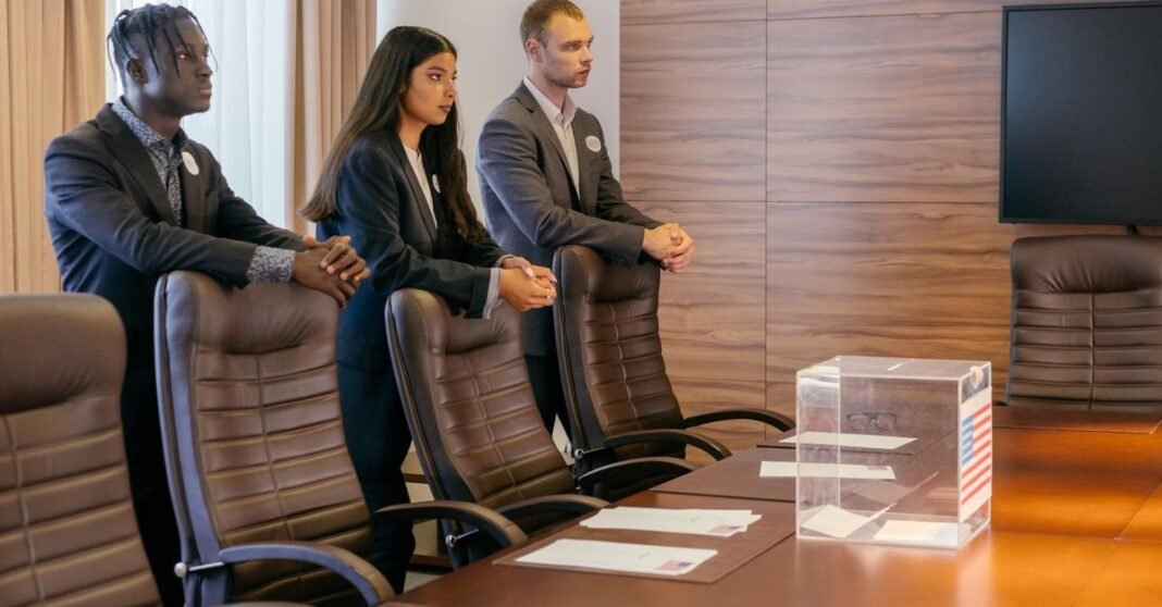 A diverse group of professionals discuss around a ballot box in a conference room.