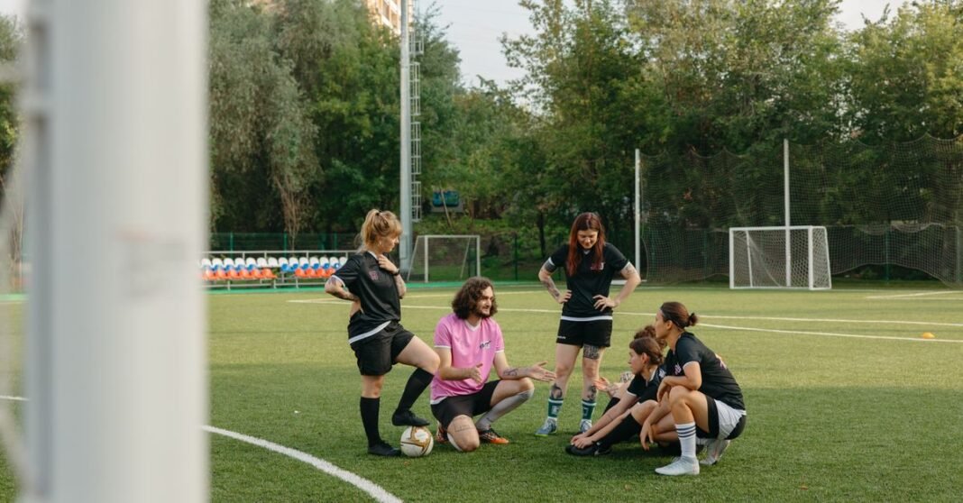 A mixed group of adults gathers on a sunny day for a team discussion on an outdoor football field.